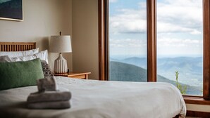 A neatly made bed with a folded towel, a bedside lamp, and a view of mountains through large windows. - Panoramic Peak: Wintergreen Condo with Views (Wintergreen Resort)