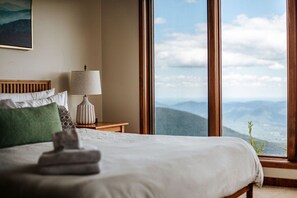 A neatly made bed with a folded towel, a bedside lamp, and a view of mountains through large windows. - Panoramic Peak: Wintergreen Condo with Views (Wintergreen Resort)