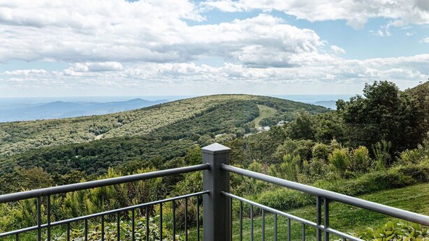 A scenic overlook with a metal railing, lush greenery, and distant mountains under a partly cloudy sky. - Panoramic Peak: Wintergreen Condo with Views (Wintergreen Resort)