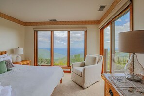 A bedroom with a large bed, a chair, a lamp, and a view of the landscape through large windows. - Panoramic Peak: Wintergreen Condo with Views (Wintergreen Resort)