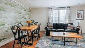 Een woonkamer met een houten eettafel, zwarte stoelen, een zwarte leren bank, een kamerkleed met een patroon, en een raam met gordijnen.