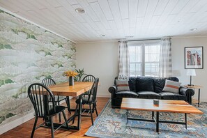 Una sala de estar con una mesa de comedor de madera, sillas negras, un sofá de cuero negro, un tapete estampado y una ventana con cortinas.