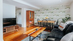 Een woonkamer met een televisie, een houten tv-meubel, een eettafel met stoelen en een bank.