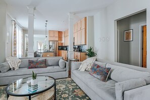 Un salon moderne avec un canapé gris, une table basse en verre et une cuisine en arrière-plan.