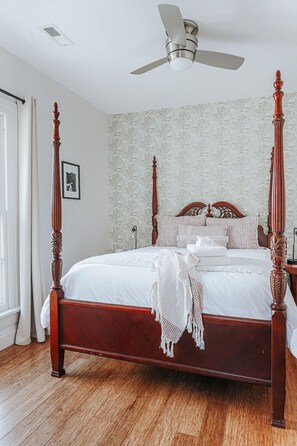 A four-poster bed with a wooden frame and white bedding, a ceiling fan, and a patterned wallpaper.