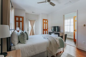 Un dormitorio con cama, ventilador de techo, televisor sobre un soporte y una puerta que da a otra habitación.