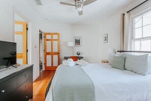 Quarto com cama, televisão, porta de madeira, escrivaninha e janela.