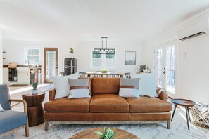 Ein modernes Wohnzimmer mit einer braunen Ledersofa, einem runden Holztisch und einem Essbereich mit weißem Tisch und Stühlen.