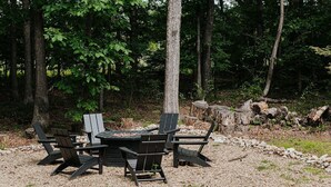 Et udendørs opholdsområde med et rundt bord og fire Adirondack-stole, omgivet af en grussti og træer.