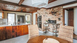 A rustic kitchen with exposed brick walls, wooden beams, and a central island with a sink and appliances. There are wicker chairs and a hanging light fixture.