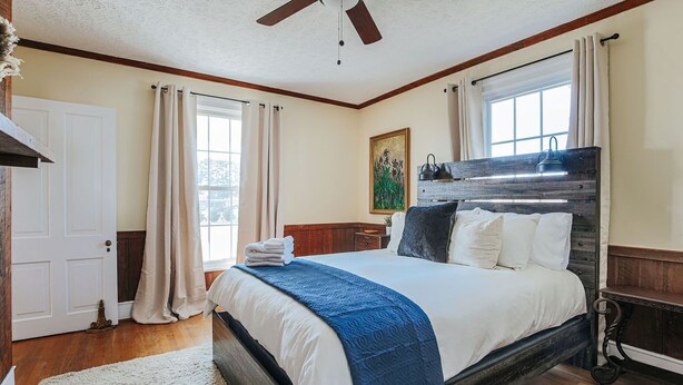 A bedroom with a bed, a ceiling fan, a window with curtains, a painting on the wall, and a wooden headboard.