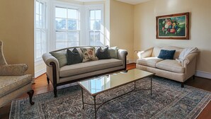 Una sala de estar con un sofá color beige, un sillón estampado, una mesa de centro de vidrio y un ventanal con marcos blancos.