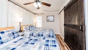 A bedroom with two beds, a ceiling fan, a window with blinds, a framed picture on the wall, and a dark wooden door. - 3 Queen Beds, 2 min to Fairgrounds, Fenced Yard (Perry)
