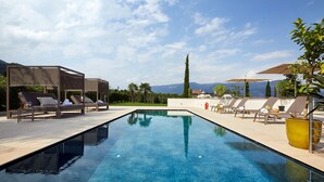 A rectangular swimming pool with clear blue water, surrounded by lounge chairs and umbrellas, set against a backdrop of mountains and trees.