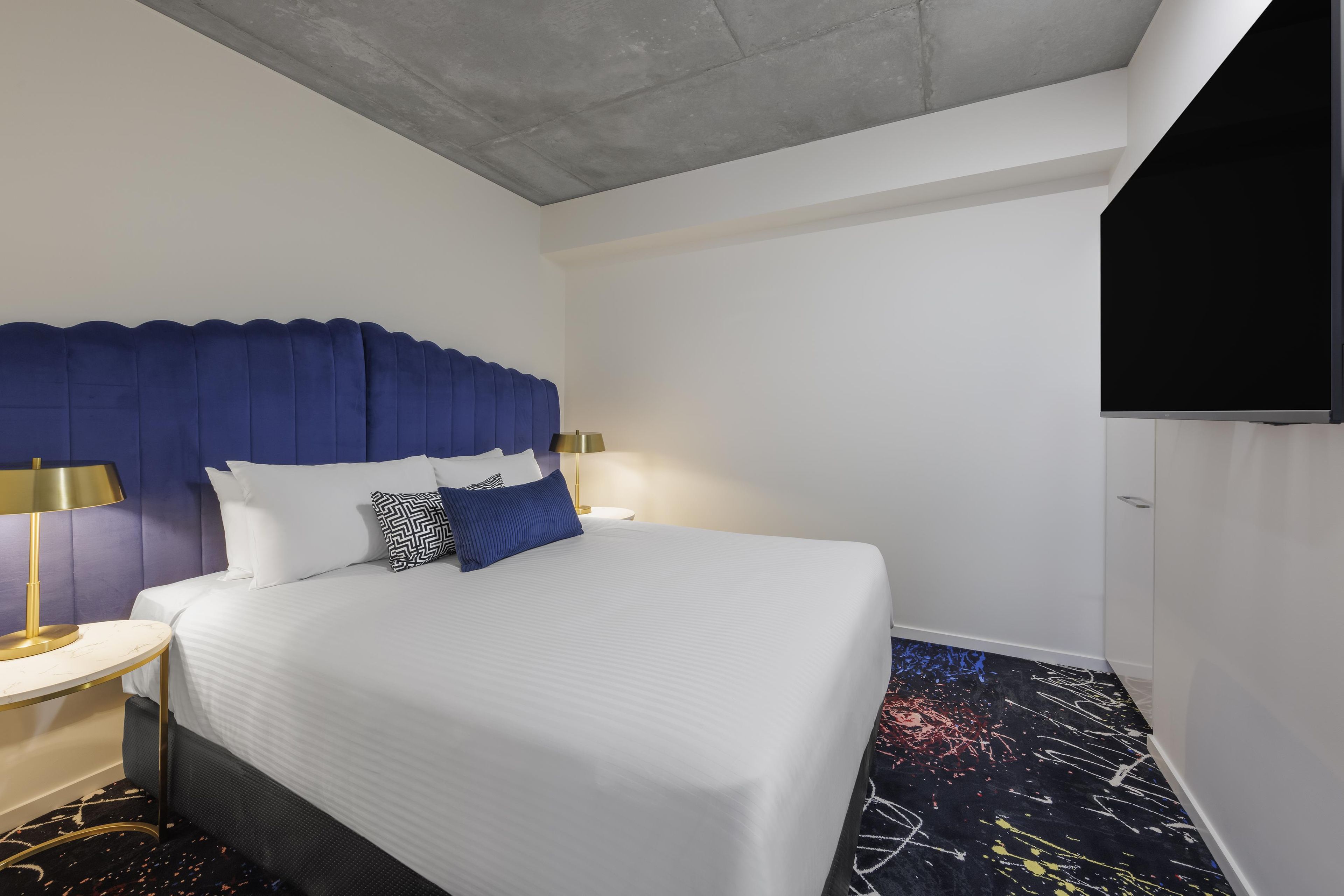 A hotel room with a large bed, a headboard, two bedside lamps, a flat-screen TV, and a patterned carpet.