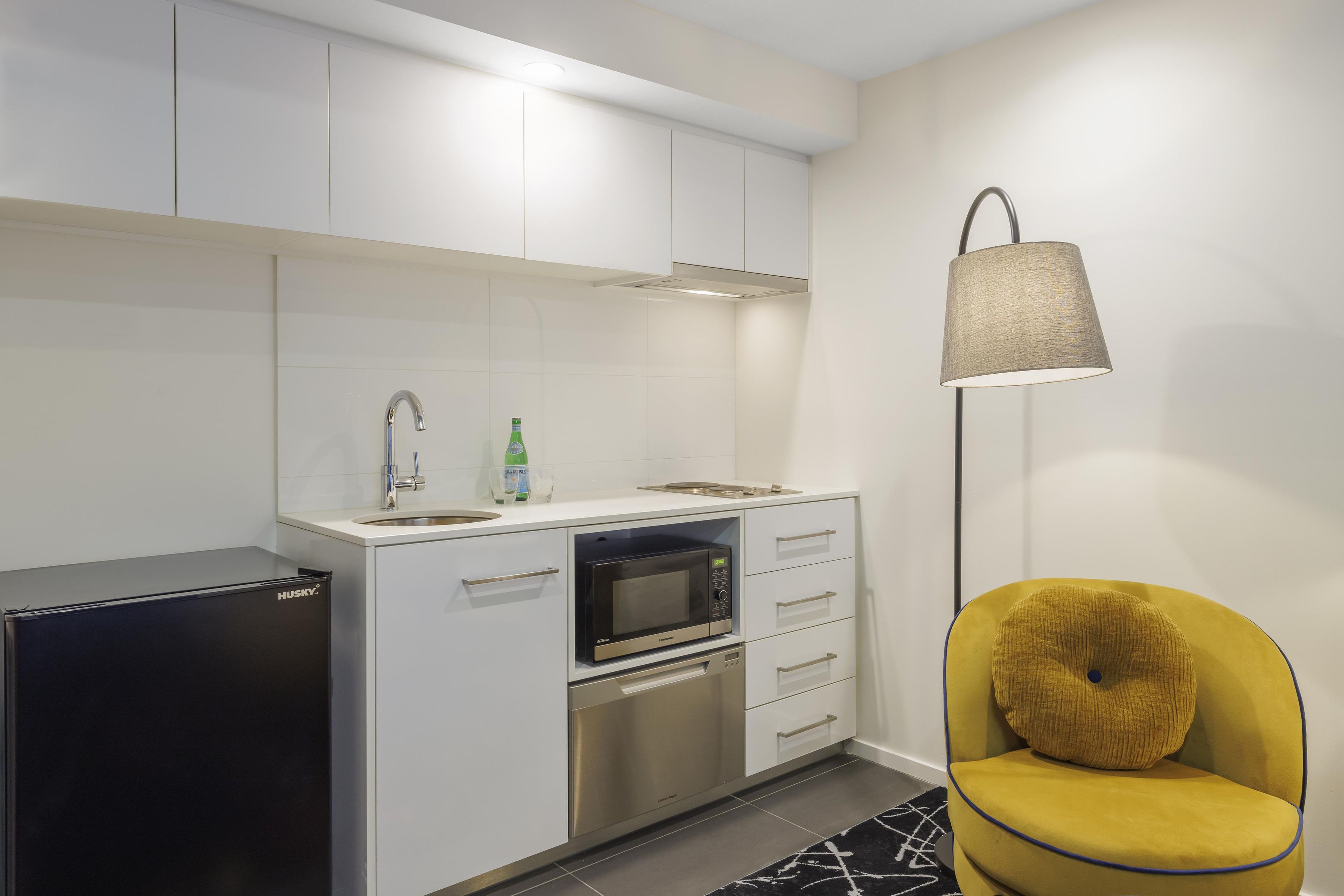Una cocina moderna con microondas integrado, horno y fregadero. Hay un refrigerador pequeño y una silla amarilla con cojín.