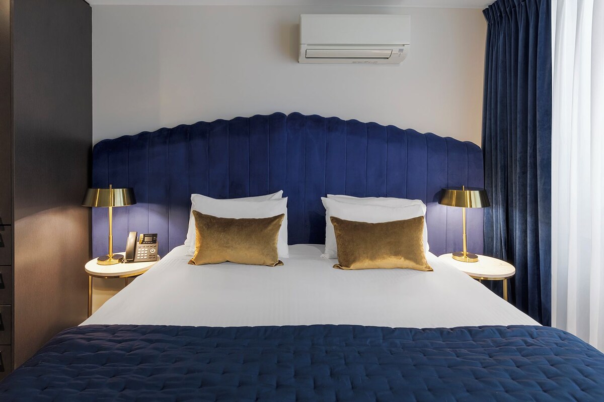 A hotel room with a large bed, two bedside tables with lamps, a wall-mounted air conditioner, and a dark blue headboard.