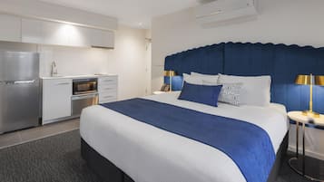 Una habitación de hotel con cama, mesita de noche, lámpara, escritorio pequeño, microondas, refrigerador y horno empotrado.