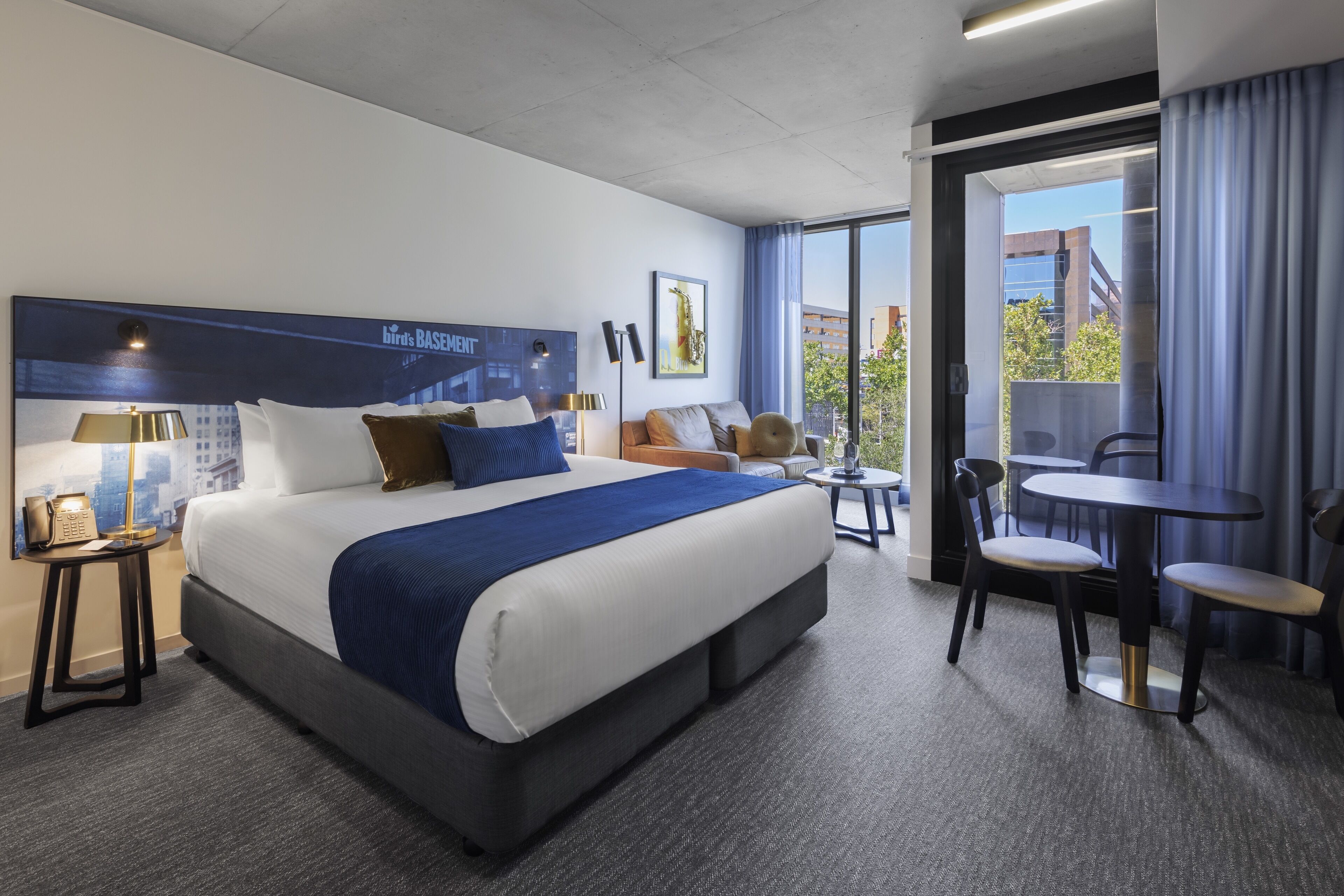 A hotel room with a large bed, a desk, and a chair. There is a window with a view of buildings and trees.