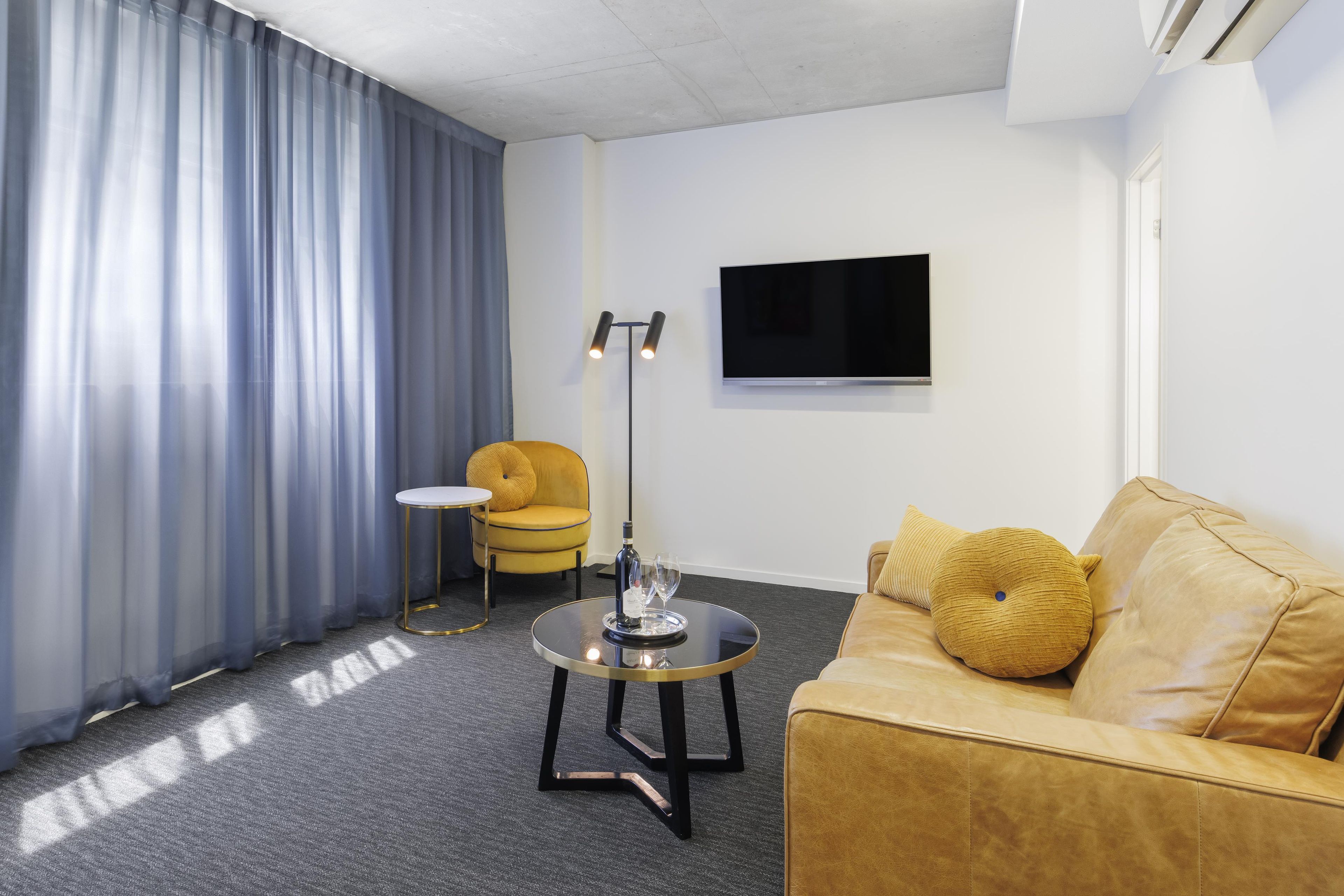 Ein modernes Hotelzimmer mit einer Couch, einem kleinen runden Tisch, einer Stehlampe und einem an der Wand befestigten Flachbildfernseher.