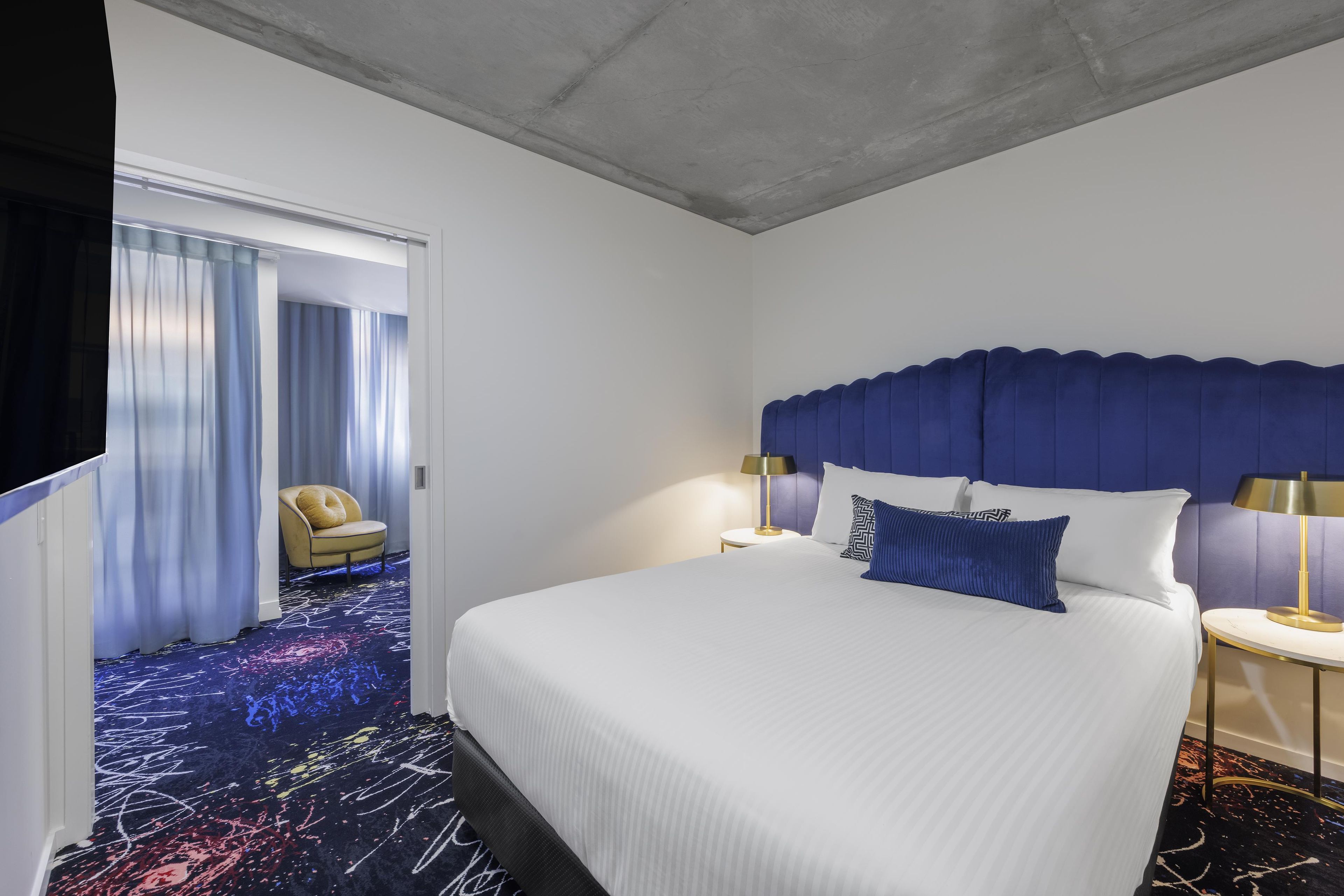 A hotel room with a large bed, two bedside tables with lamps, a chair, and a view of a hallway with curtains.
