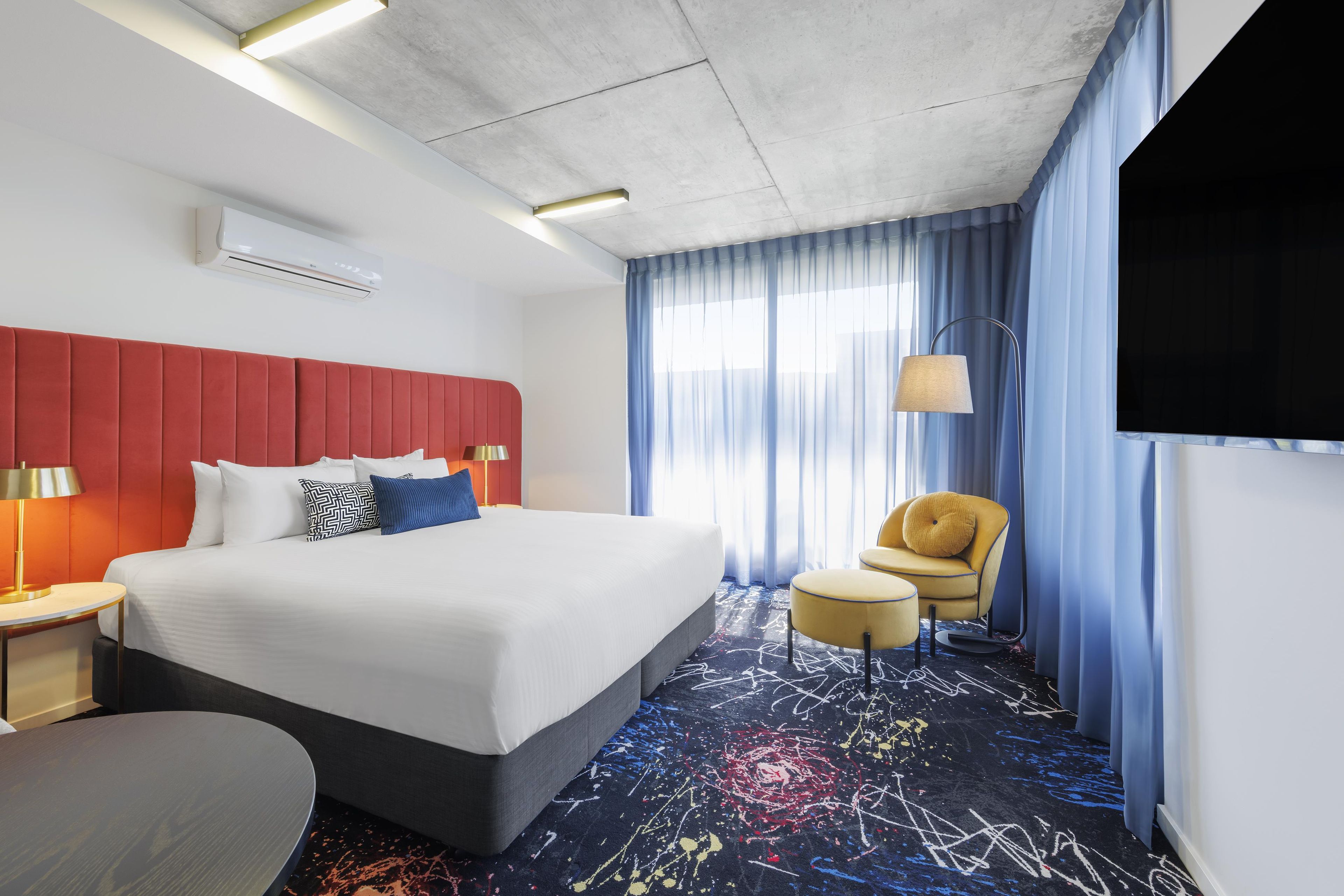 Quarto de hotel com uma cama grande, cabeceira vermelha, televisão de tela plana, escrivaninha com luminária e uma cadeira.