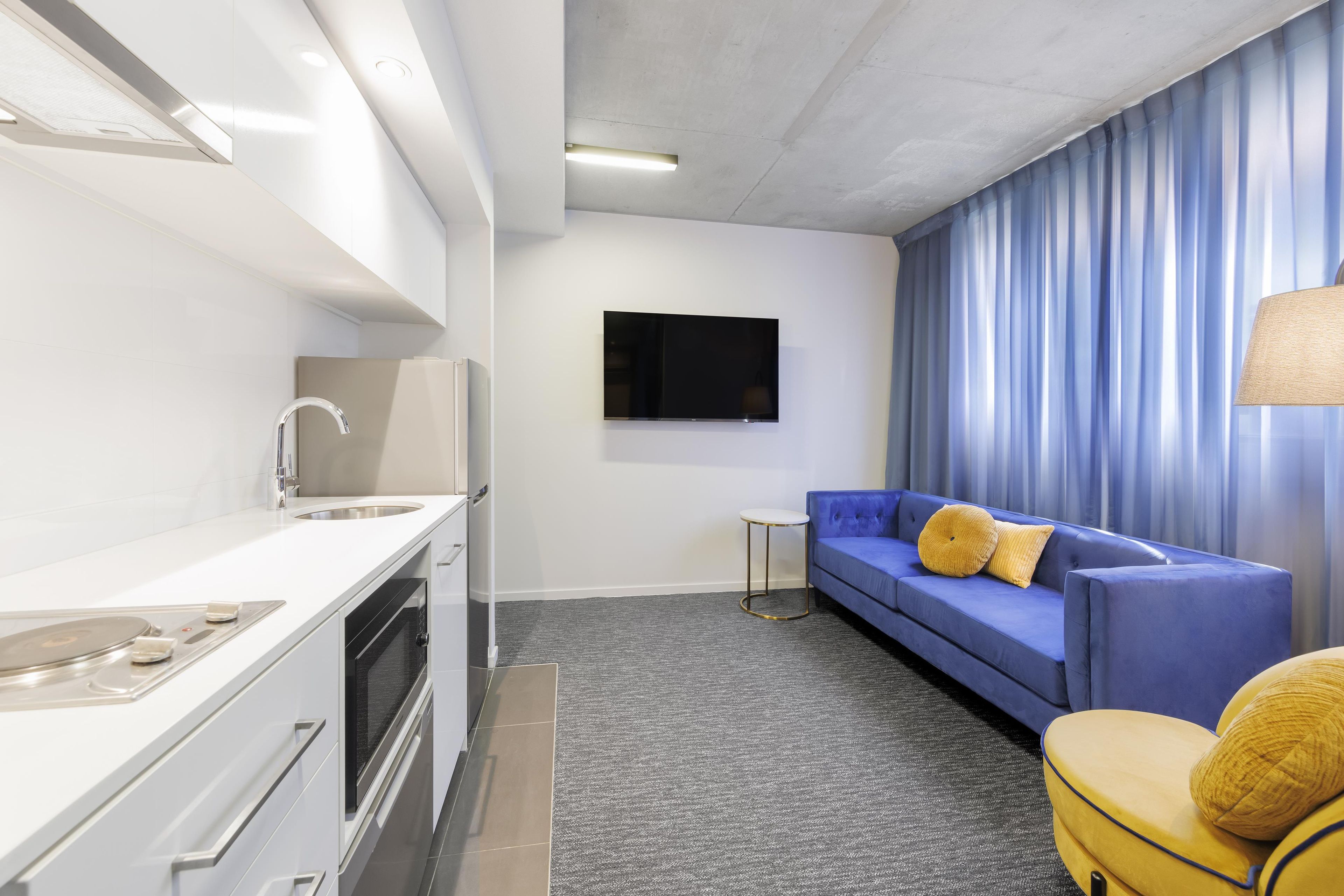 Une chambre d’hôtel moderne équipée d’une kitchenette, d’un téléviseur à écran plat fixé au mur, d’un canapé bleu avec des coussins jaunes et d’une petite table ronde.