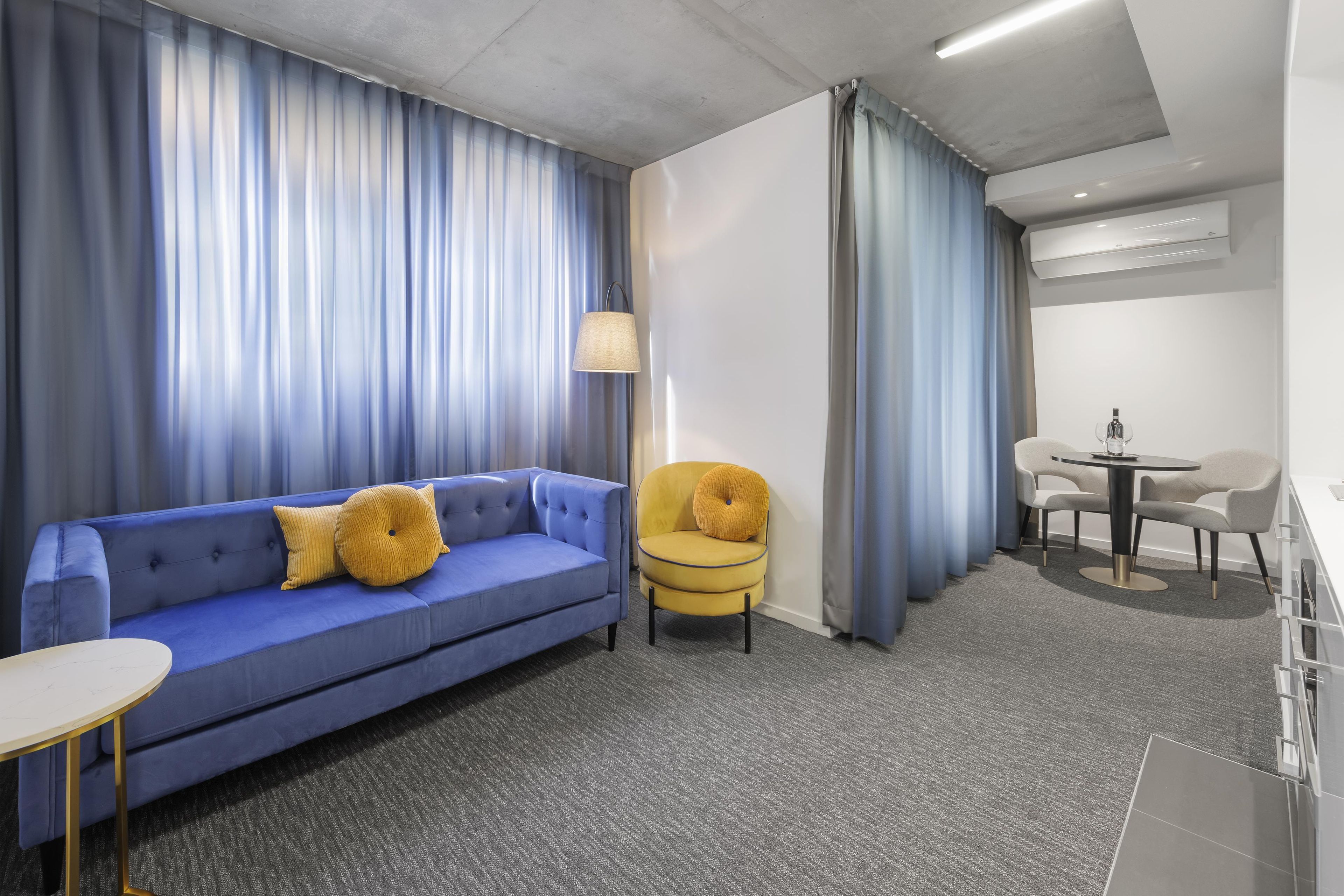 Een hotelkamer met een blauwe bank, een gele fauteuil, een kleine tafel en een bureau met stoelen.