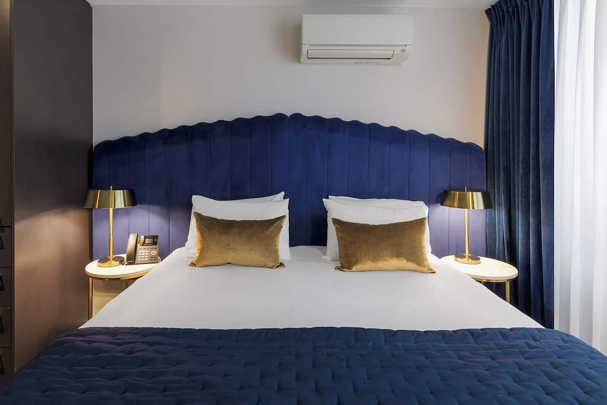 A hotel room with a large bed, two bedside tables with lamps, a wall-mounted air conditioner, and a dark blue headboard.