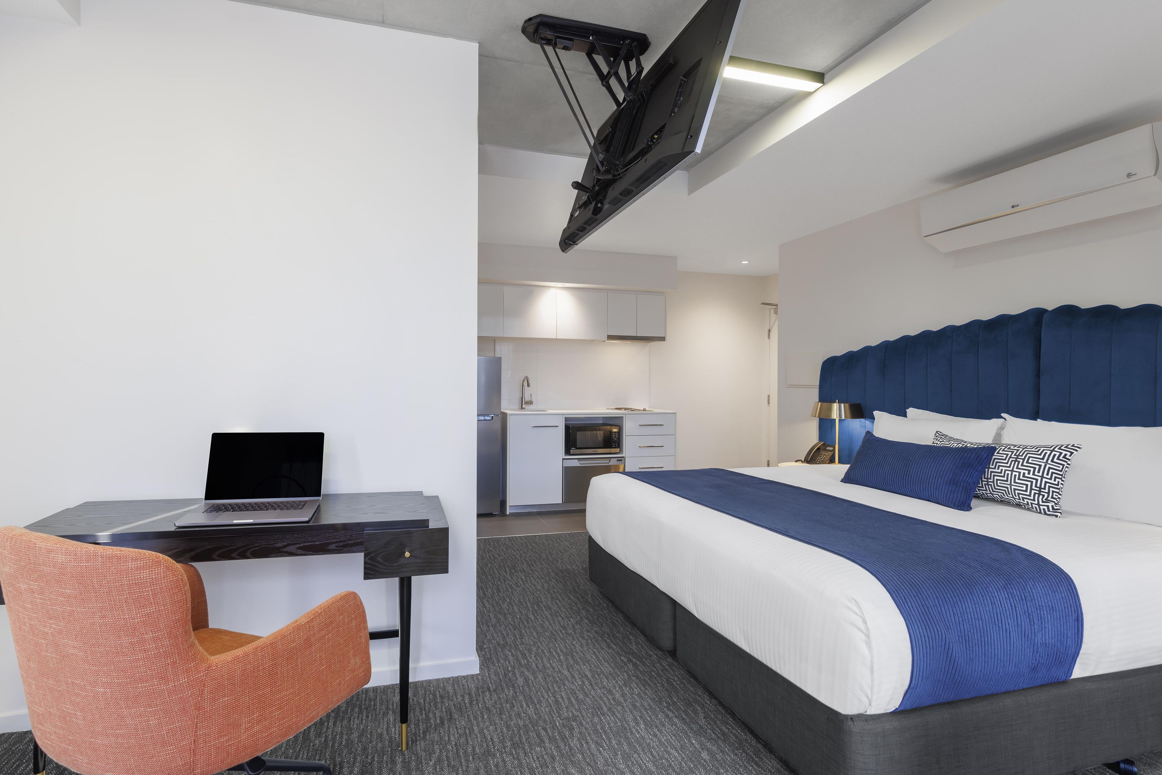 A modern hotel room with a bed, desk, chair, kitchenette, and a ceiling-mounted projector.