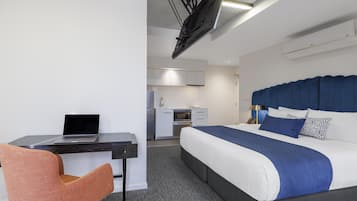 A modern hotel room with a bed, desk, chair, kitchenette, and a ceiling-mounted projector.