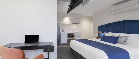 A modern hotel room with a bed, desk, chair, kitchenette, and a ceiling-mounted projector.