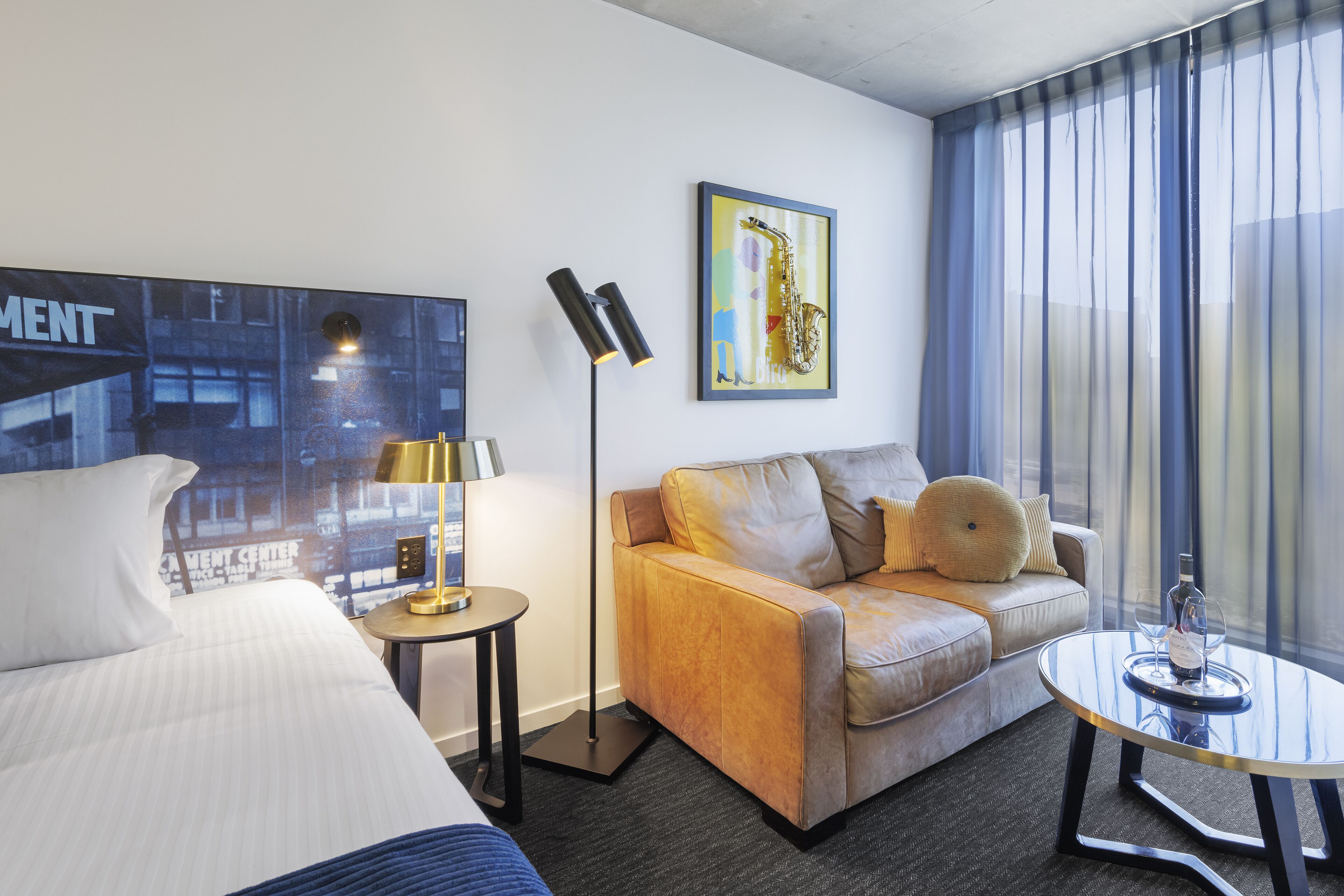 A hotel room with a bed, a sofa, a small table, a floor lamp, and a painting of a saxophone.