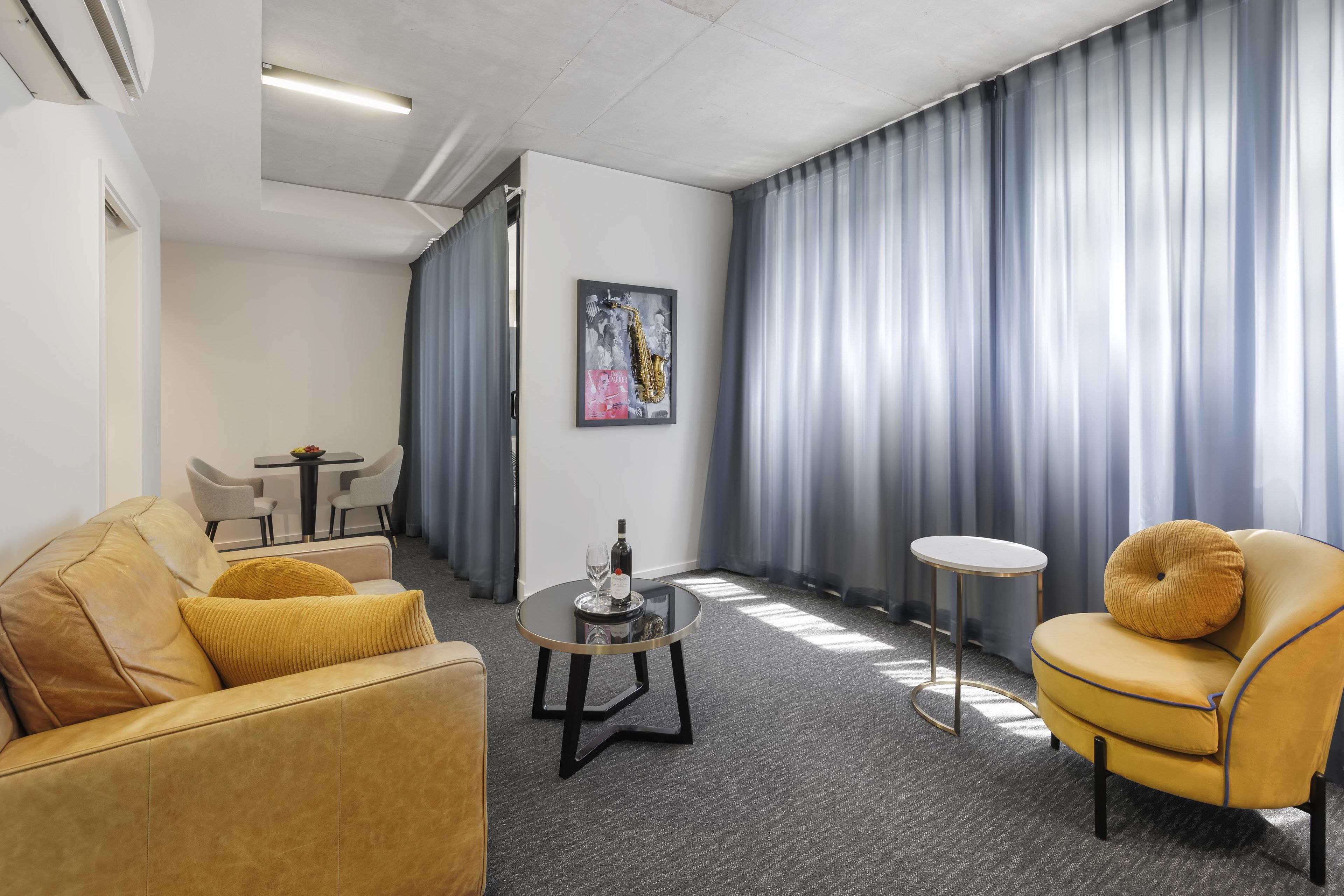 A modern hotel room with a leather sofa, a yellow armchair, a small round table with a bottle and glass, and a painting on the wall.
