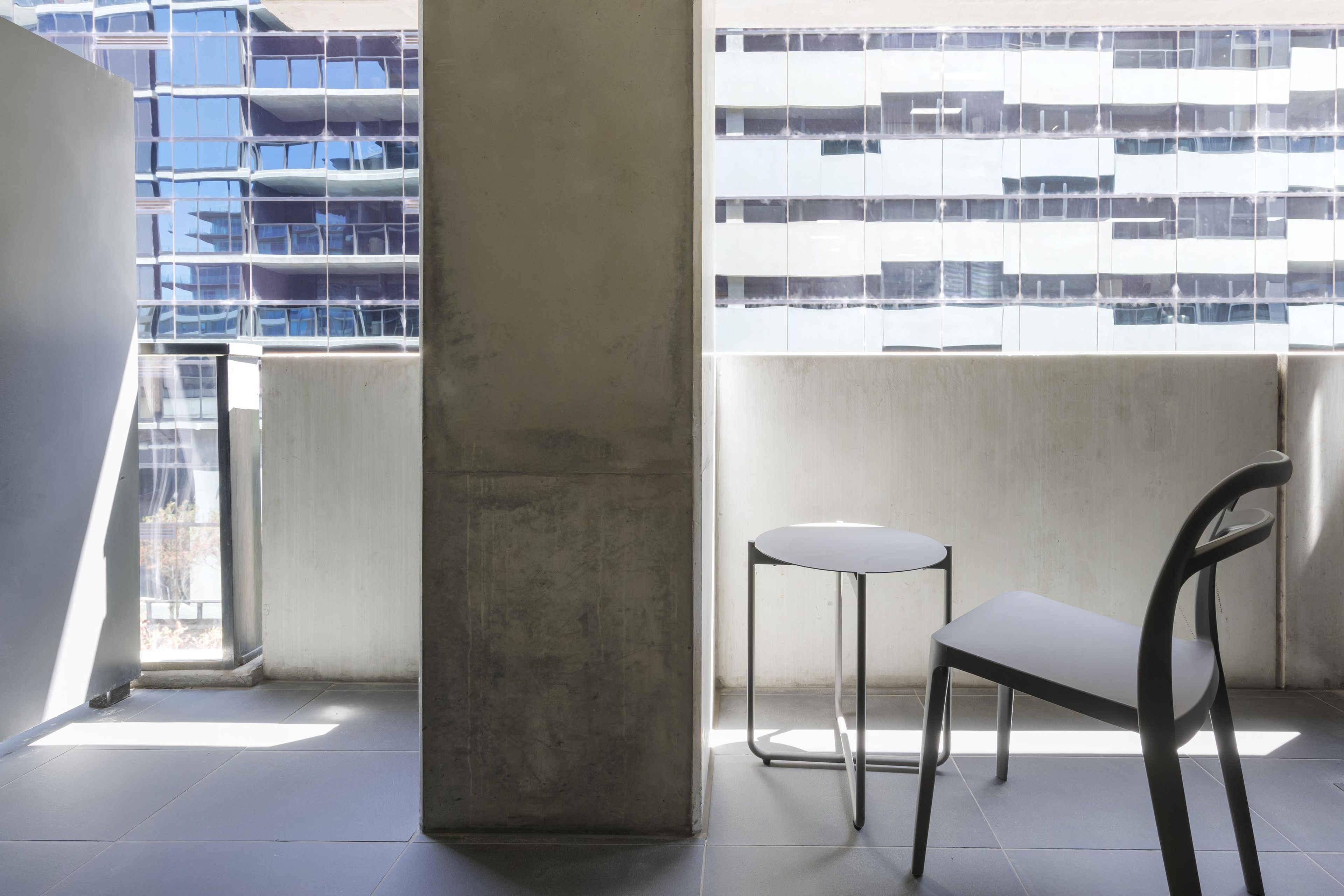 A balcony with a chair and a small table, a concrete pillar, and a view of a modern building with reflective windows.