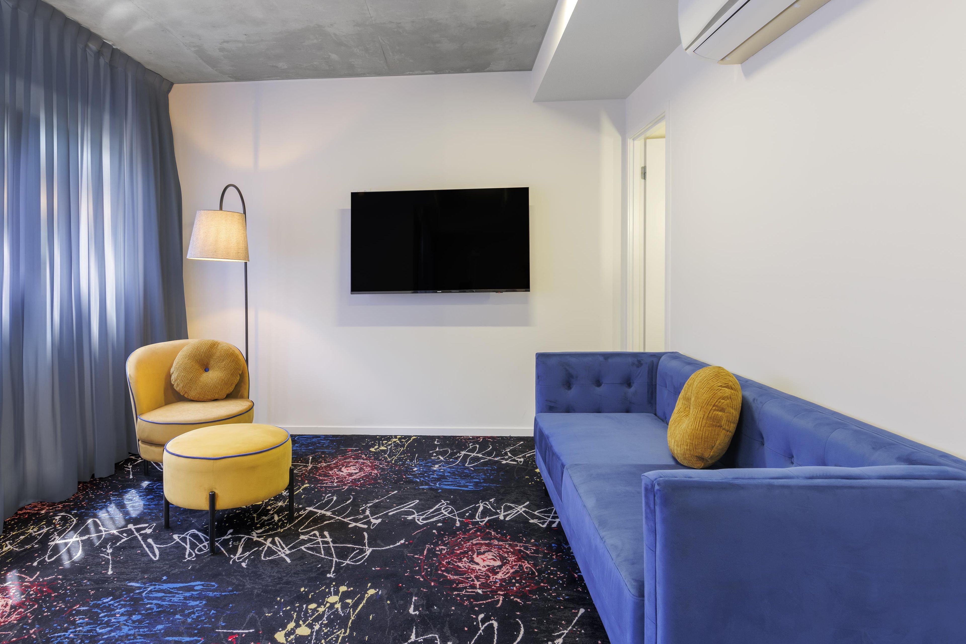 Een moderne woonkamer met een flatscreen televisie aan de muur, een gele fauteuil met een rond kussen, een blauwe bank met een rond kussen, en een kamerbreed tapijt met een patroon.