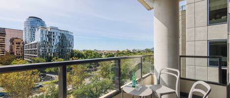 A balcony with a table and chairs, overlooking a cityscape with modern buildings and a clear sky.