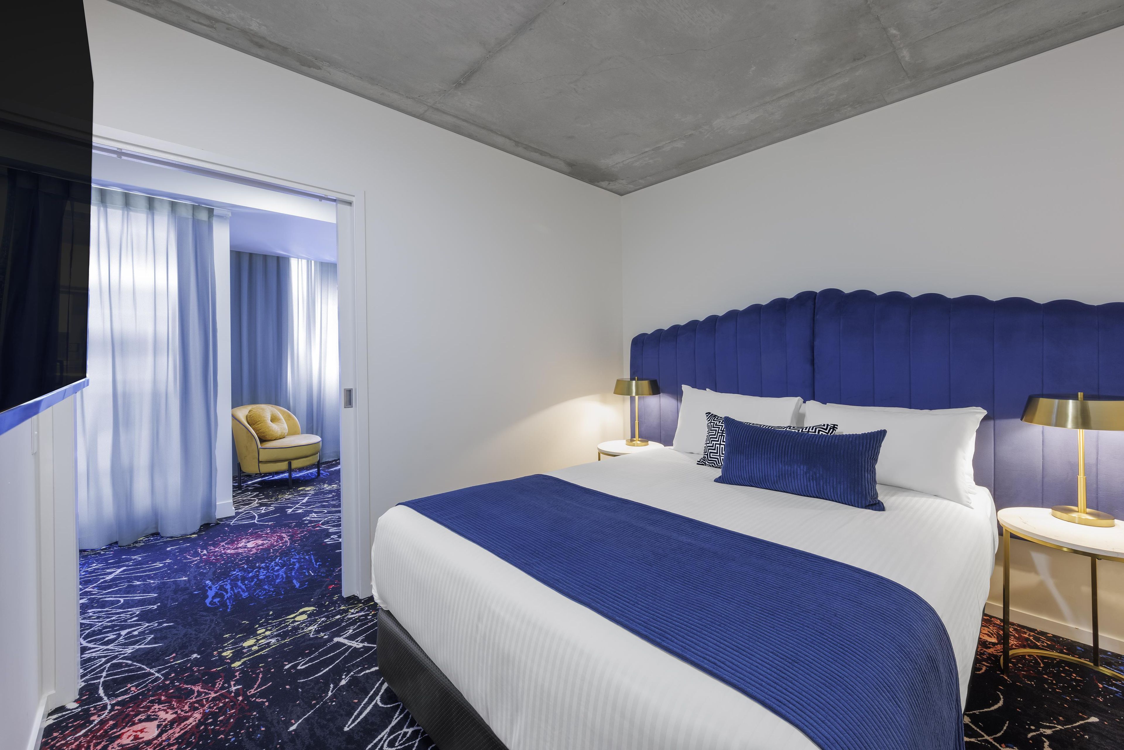 Habitación de hotel con una cama grande, una silla, una mesita de noche y vistas a un pasillo con una puerta.