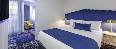 Habitación de hotel con una cama grande, una silla, una mesita de noche y vistas a un pasillo con una puerta.
