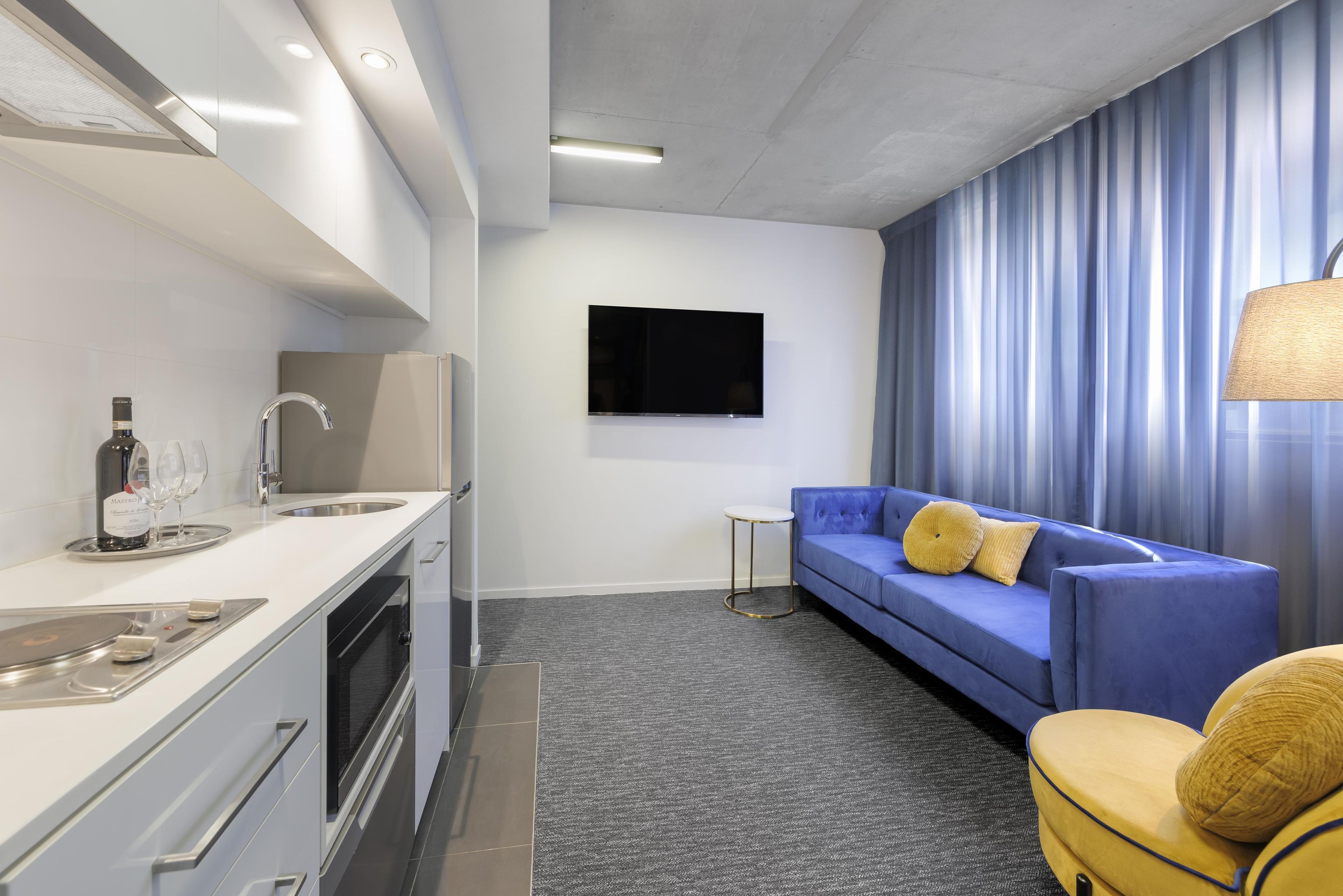 A modern hotel room with a kitchenette, a flat-screen TV mounted on the wall, a blue sofa with yellow cushions, and a small side table.