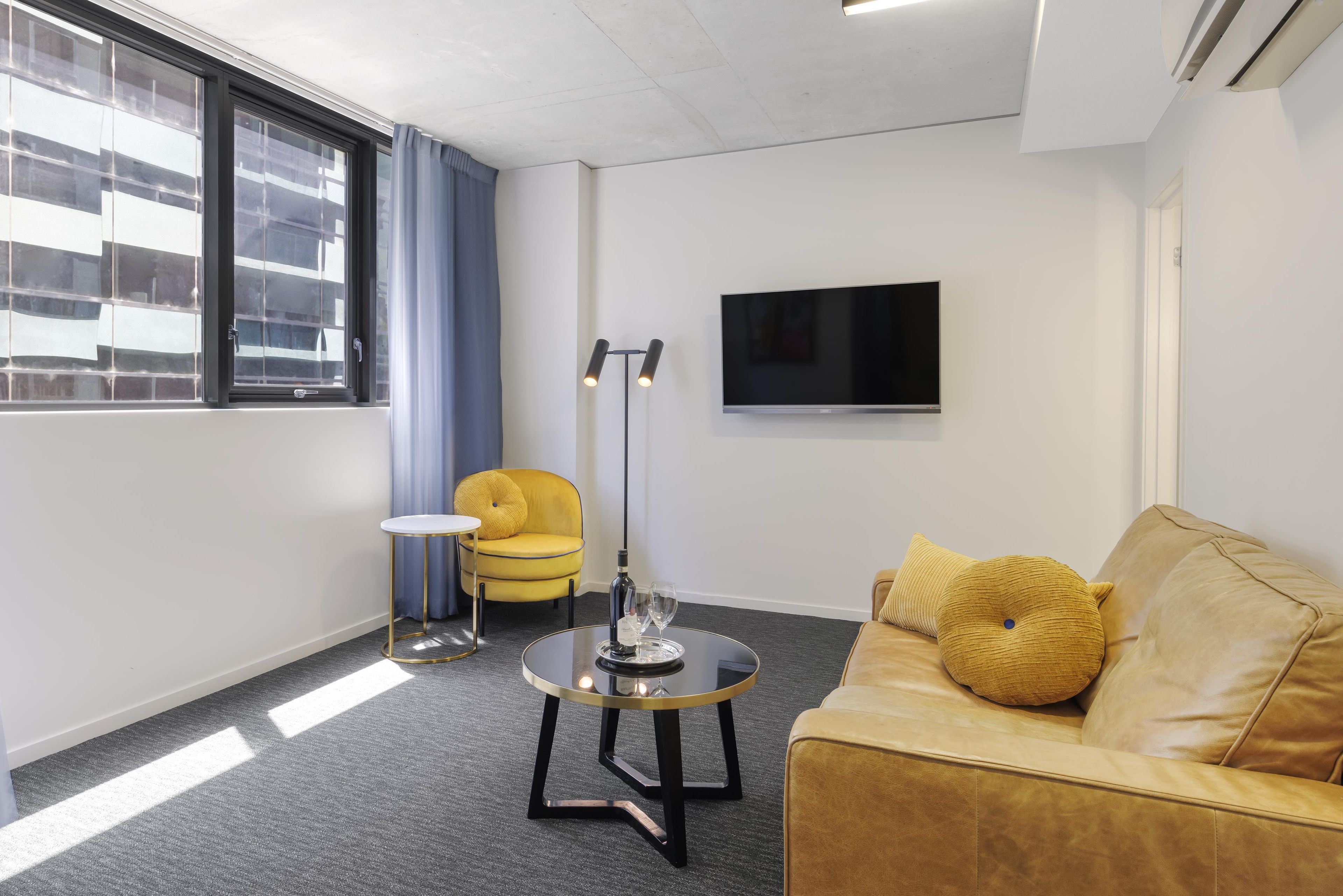 Een moderne woonkamer met een bank, een klein rond tafeltje, een stoel, een aan de muur bevestigde televisie en een raam met gordijnen.