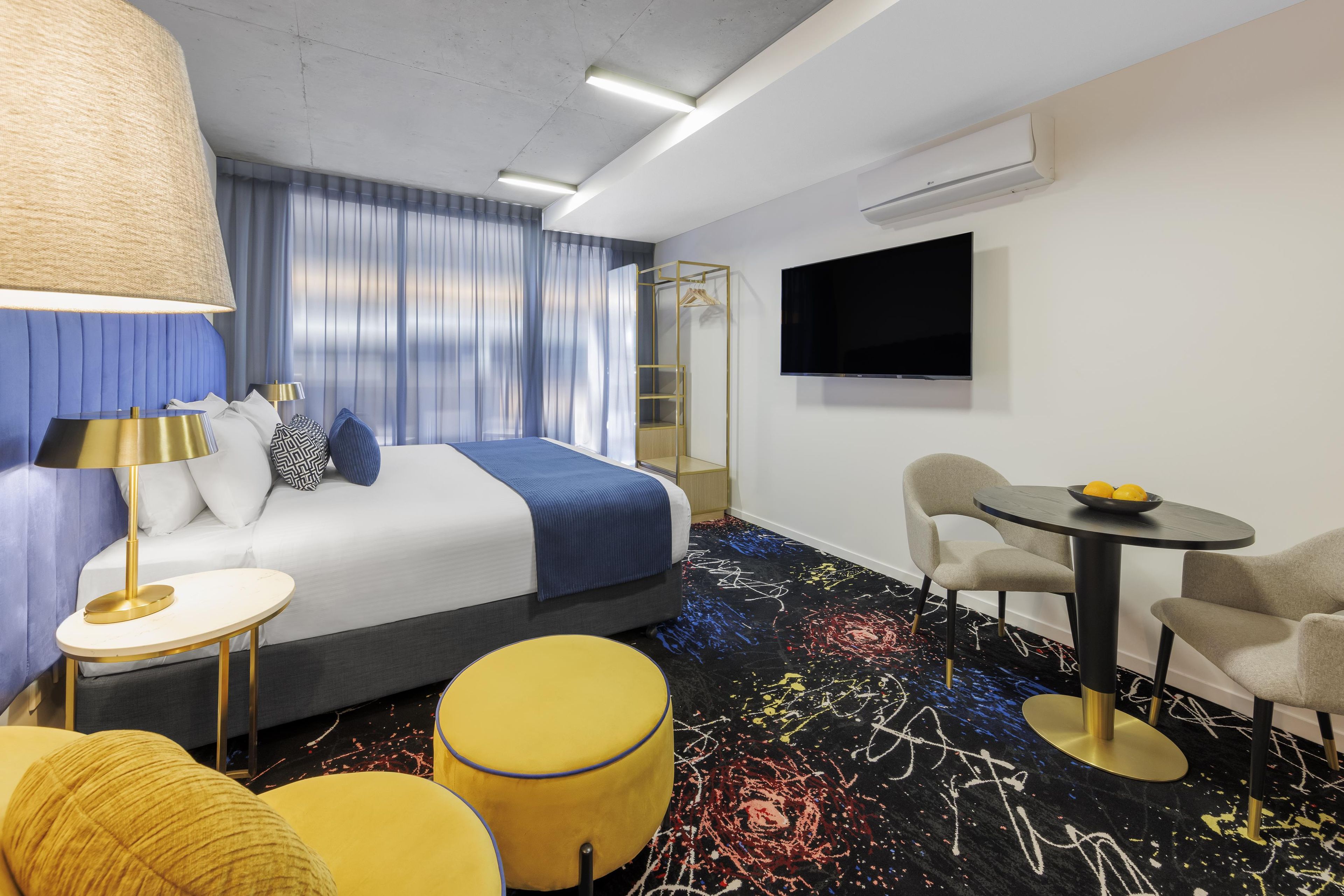 Habitación de hotel con cama, mesita de noche, lámpara, sillas, televisor y una pequeña mesa con fruta.