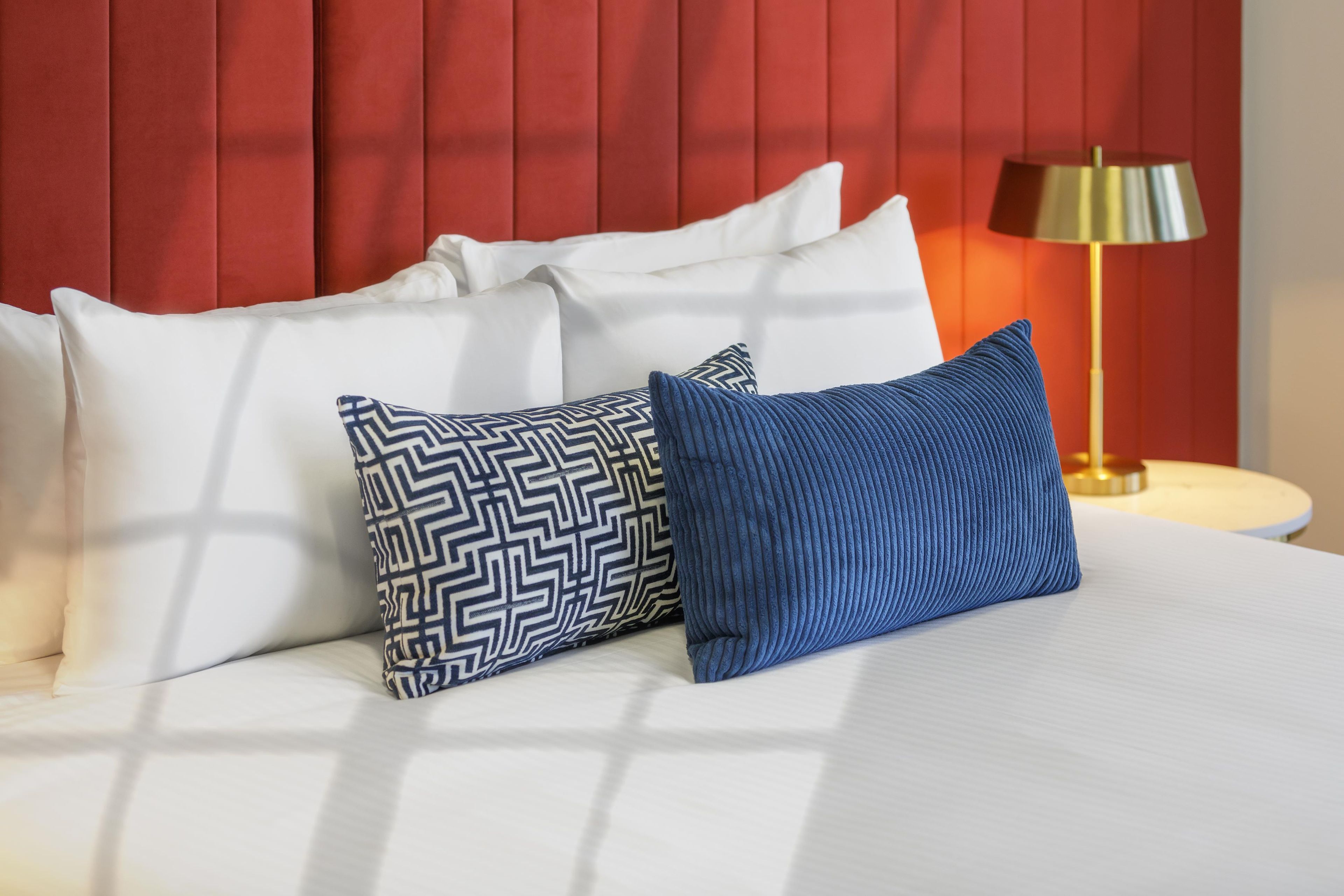 A bed with white bedding and two patterned pillows, a red headboard, and a lamp on a bedside table.