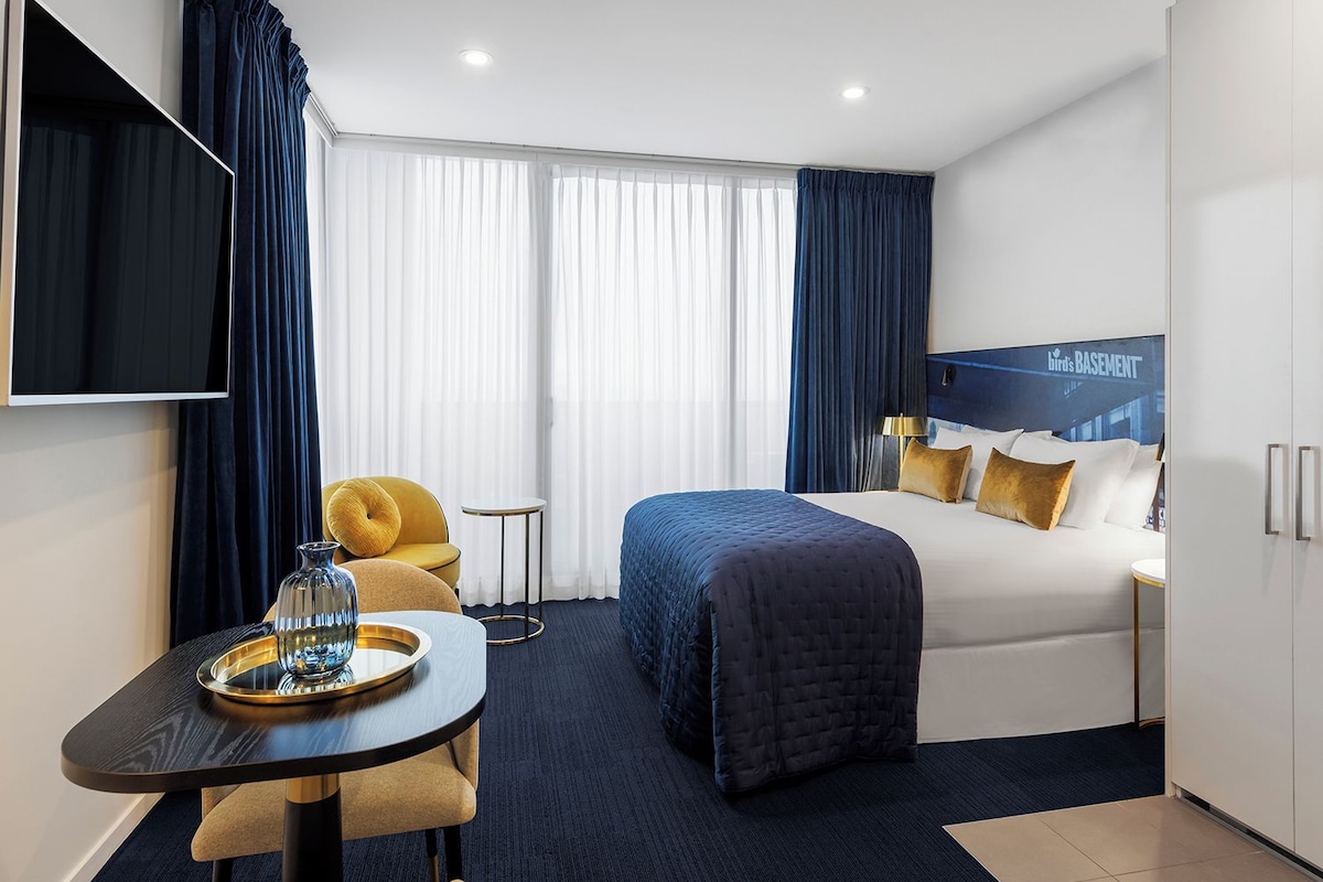 A hotel room with a bed, a desk with a chair, a TV, and a view of the sky through the window.