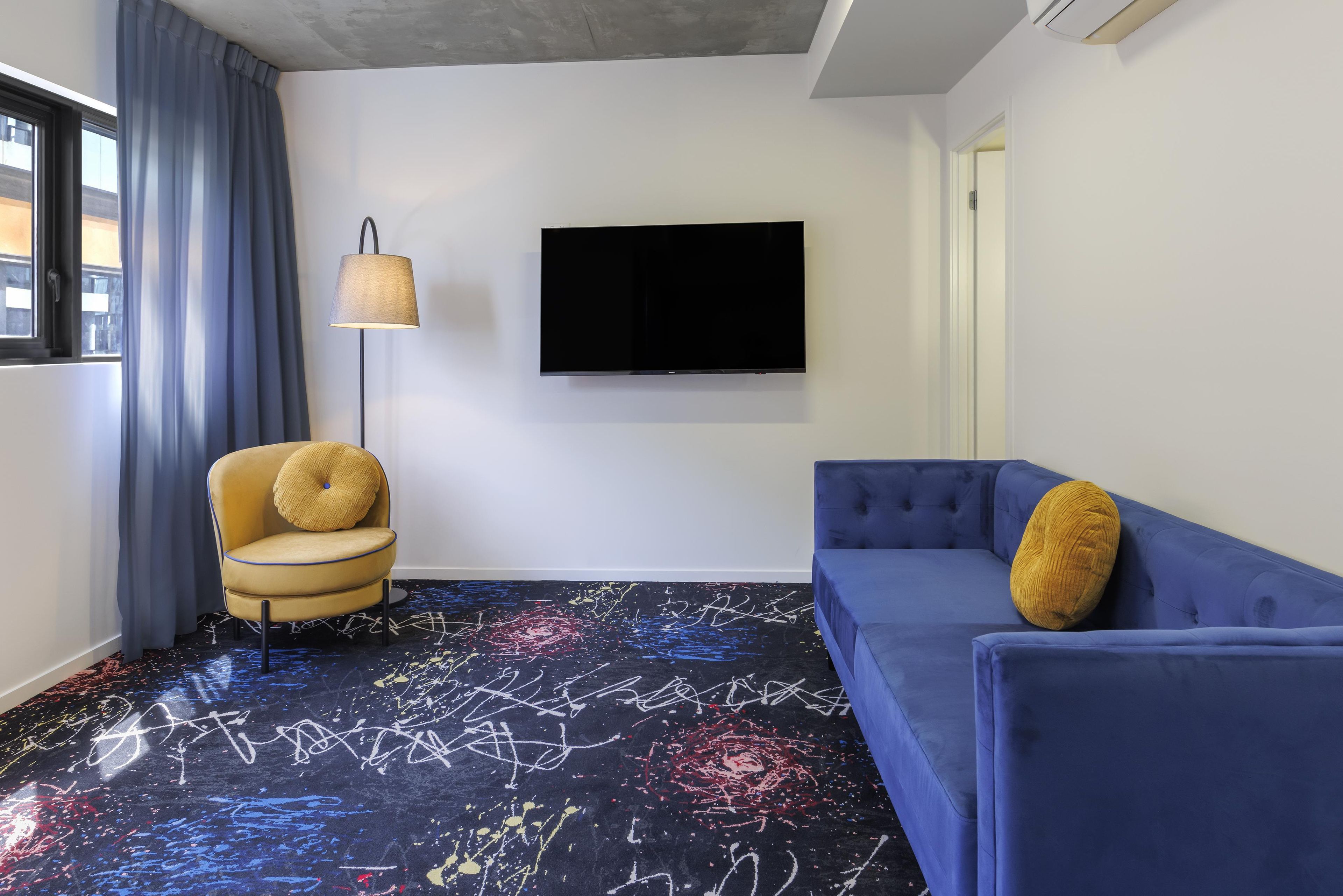 Una habitación con un sofá azul, un sillón amarillo, una lámpara de pie y un televisor de pantalla plana montado en la pared.
