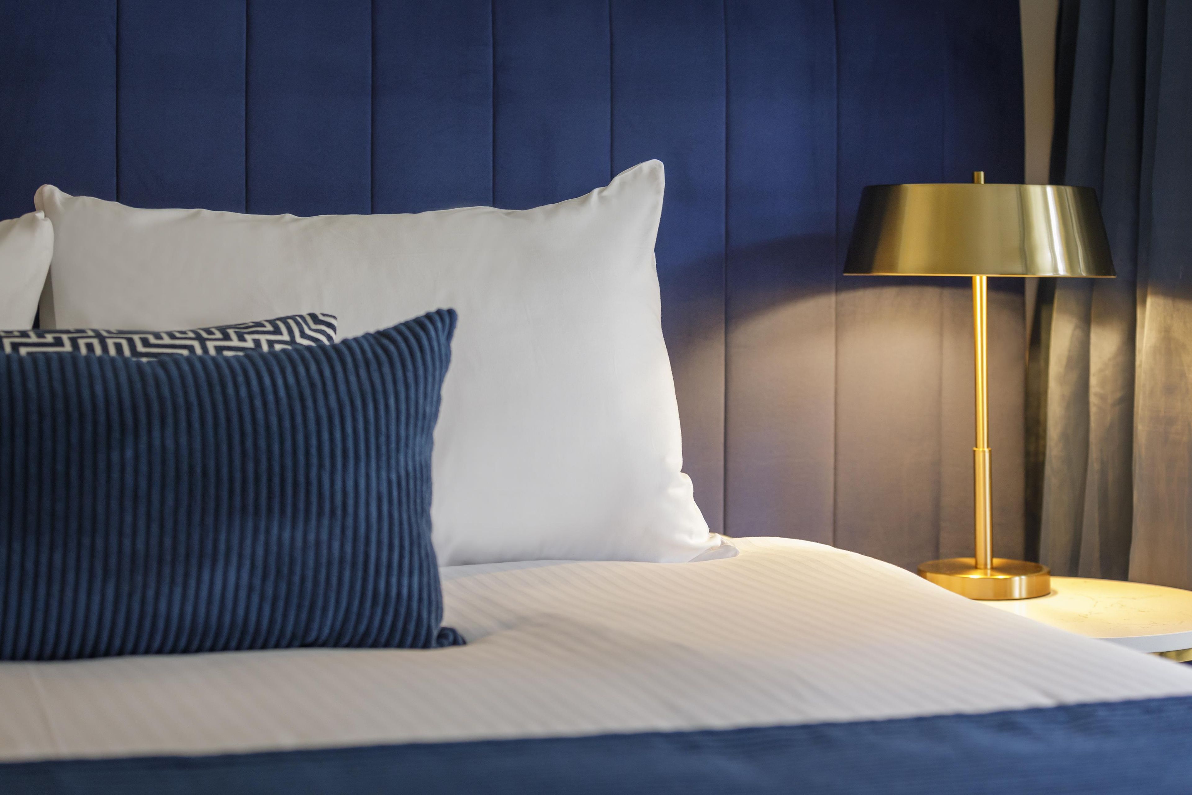 Una cama con ropa blanca y dos almohadas, una lisa y otra con un diseño geométrico, frente a una pared azul.