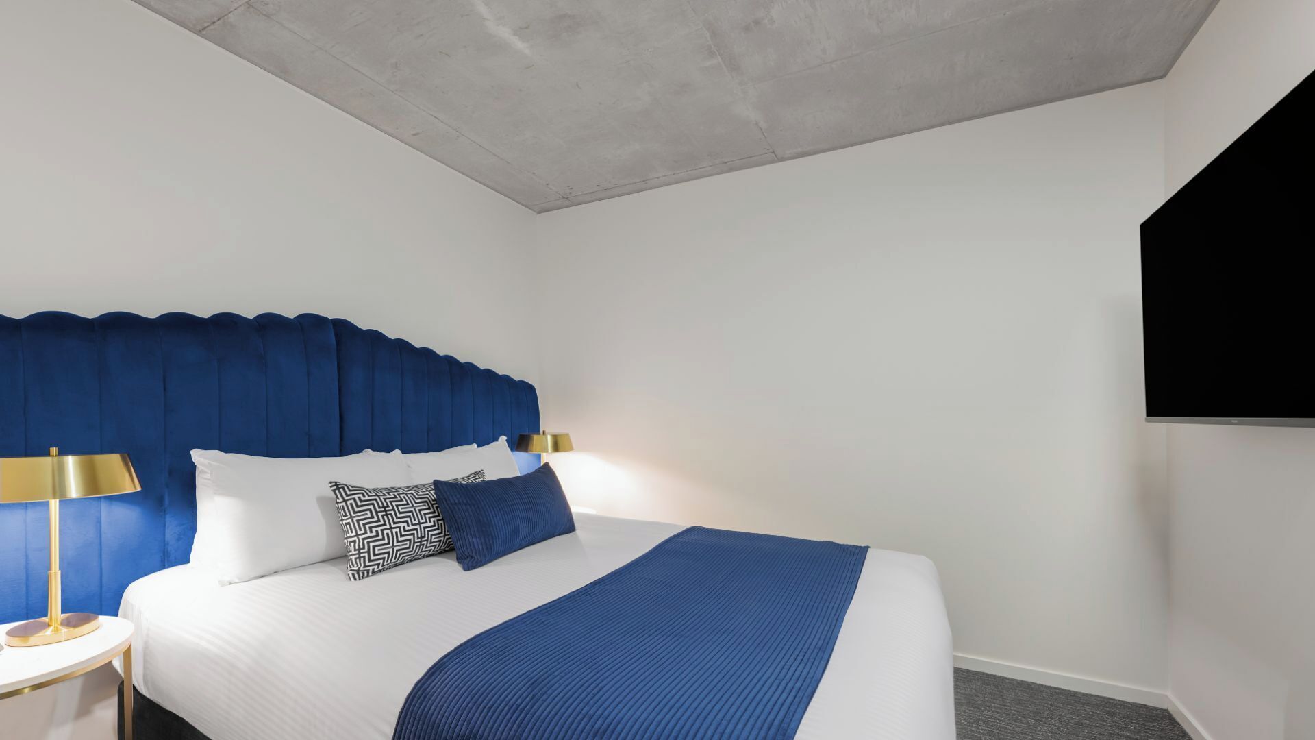 Una habitación de hotel con una cama grande, un cabecero azul, una lámpara dorada y un televisor negro en la pared.