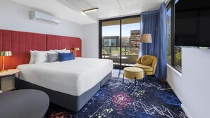 Ein modernes Hotelzimmer mit einem großen Bett, einem roten Kopfteil, einem runden Tisch, einem Sessel und Blick auf die Stadt durch große Fenster.