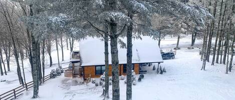 A cabin surrounded by snow-covered trees, with a wooden fence and a car parked nearby.