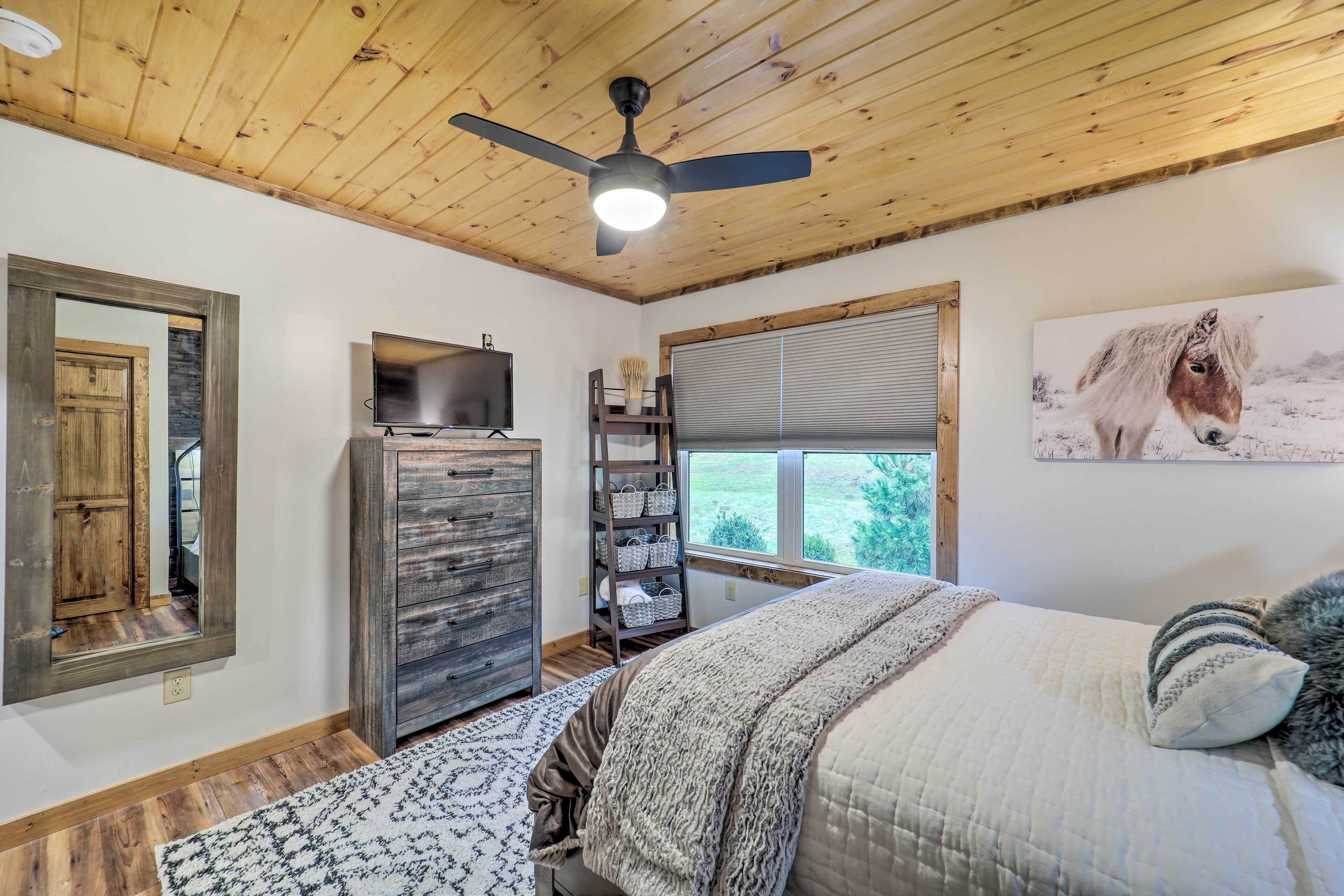 Une chambre avec un plafond en bois, un ventilateur de plafond, un lit recouvert d’une couverture à motifs, une commode, un téléviseur, une fenêtre avec des stores et un tableau représentant un cheval.